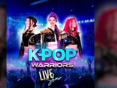 KPOP Warriors promotional artwork featuring three costumed performers with colorful hair on a bright blue stage, with the KPOP Warriors logo displayed prominently in front.
