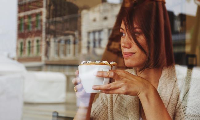 Photo from outside of window of a woman drinking coffee with reflection of buildings showing