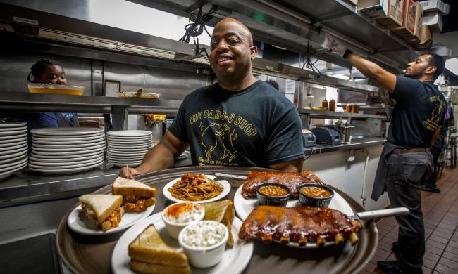 Owner Eric Vernon holds large serving tray with slabs of rib, bbq spaghetti and pulled pork sandwiches